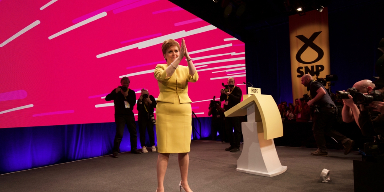 Nicola Sturgeon stood on stage at a SNP conference. She is holding the palms of her hands together at face level and wears a yellow jacket and skirt. In the background are several photographers, a pink screen and SNP logo
